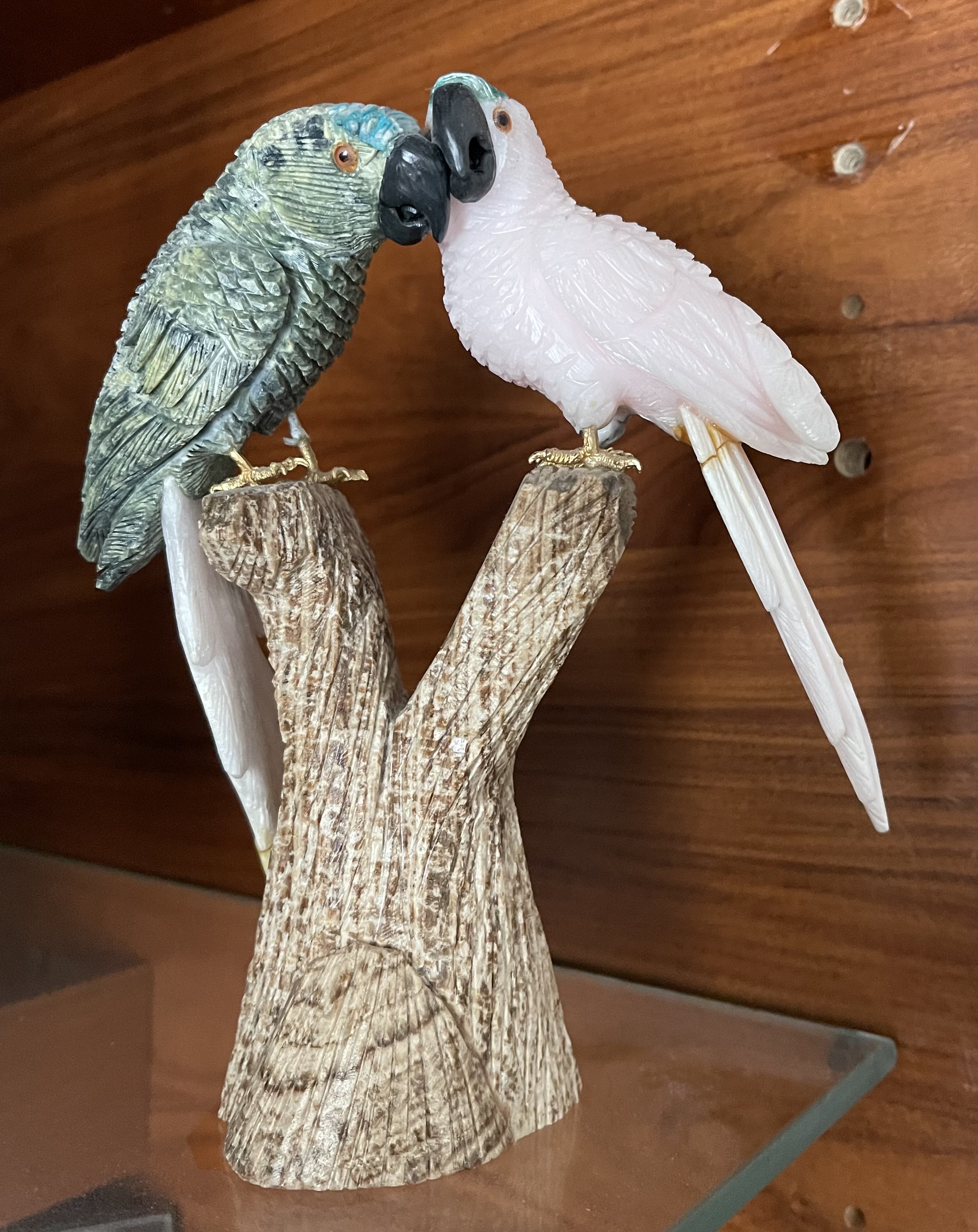 Hand-carved memento from Iguazu Falls — two stone parrots, one green-blue and one rose-white, perched on a wooden branch.