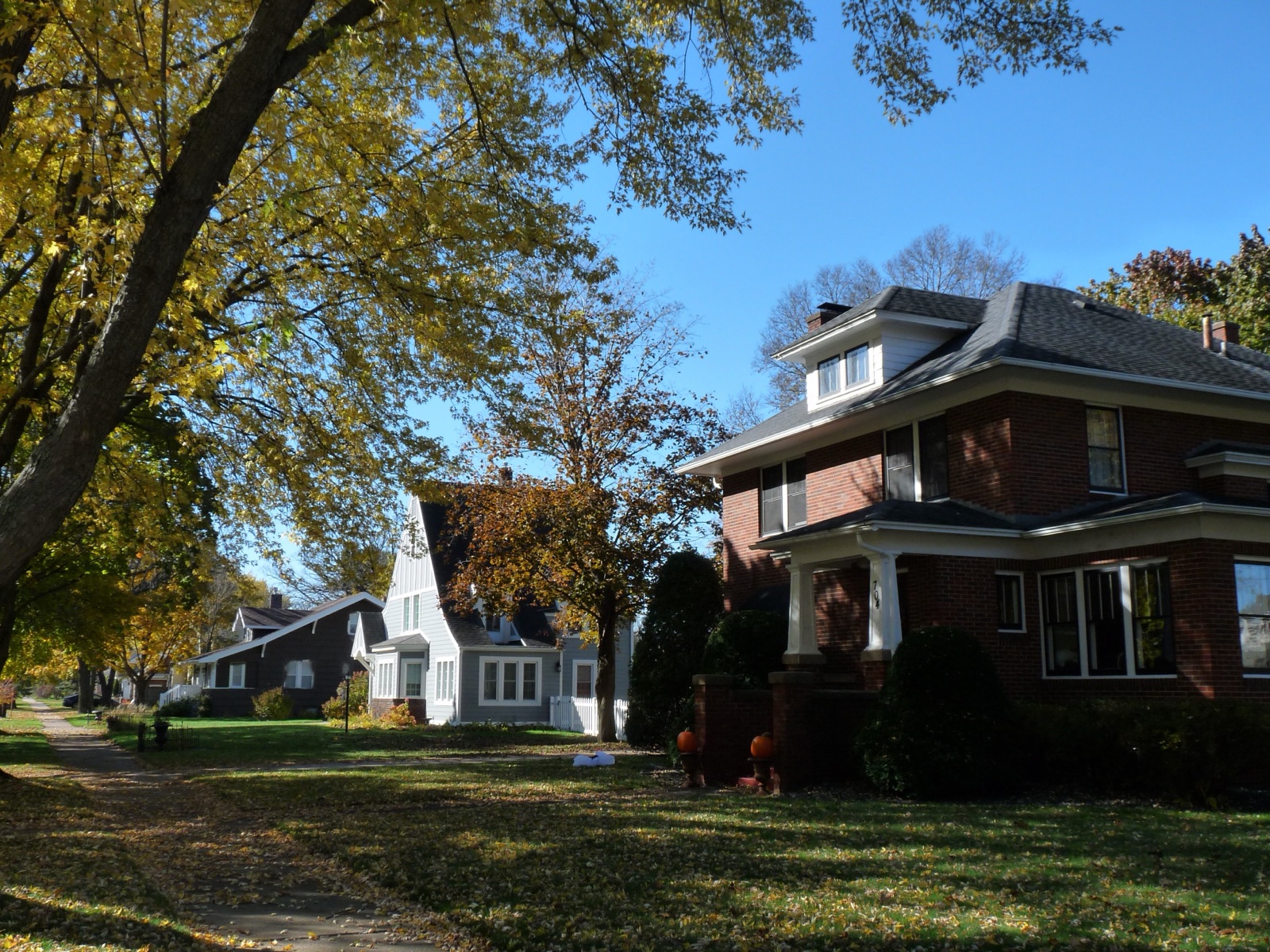 A brick American Foursquare with a deep white-columned porch on a tree-lined street in autumn, photographed from the sidewalk.