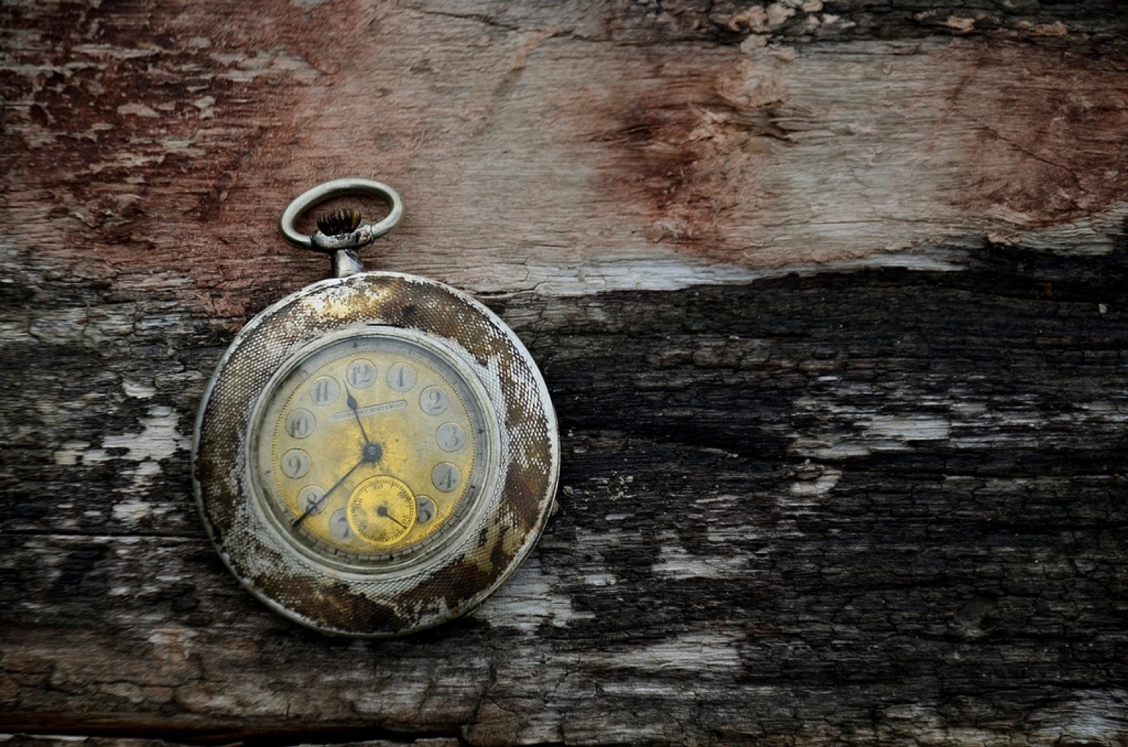 An antique pocket watch, its case worn to a mottled patina, lying face-up on weathered gray barn wood.